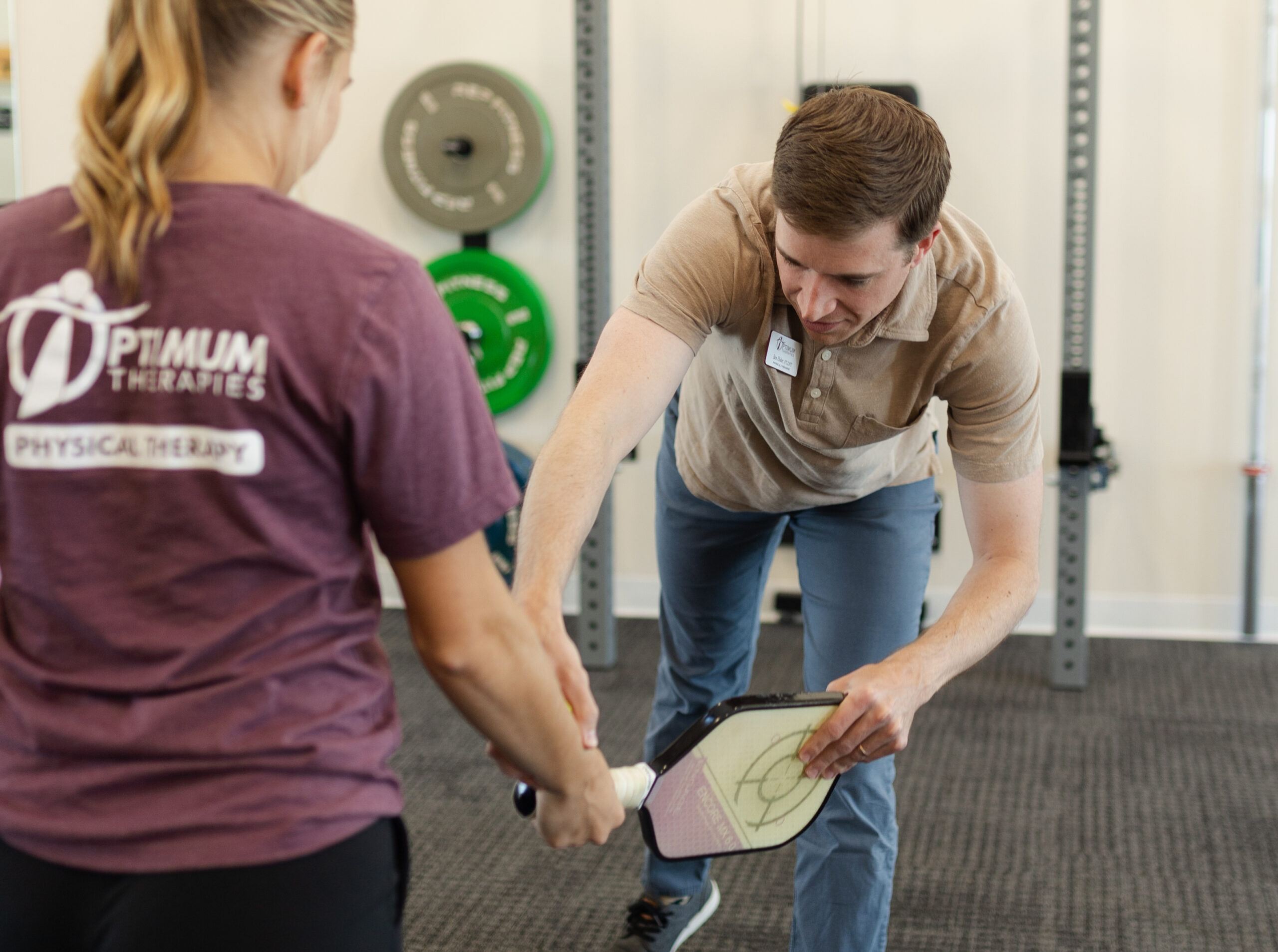 A physical therapist guides a woman holding a pickleball paddle, helping her with proper technique in a therapy clinic setting.