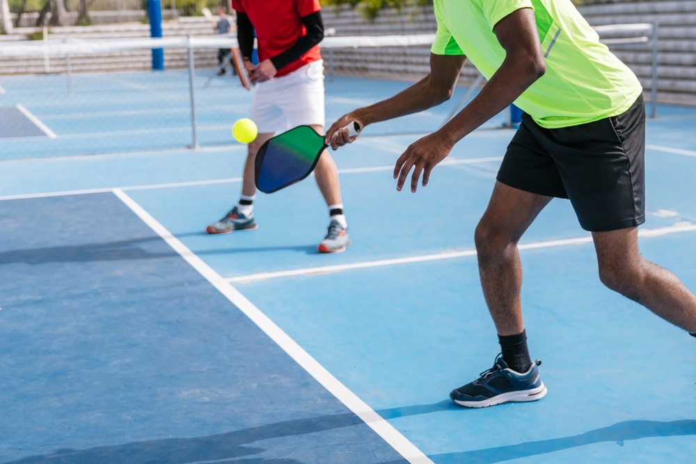 Two people playing pickleball on an outdoor blue court. One player in a neon shirt and black shorts is about to hit a yellow ball with a paddle, while the other stands ready in the background.