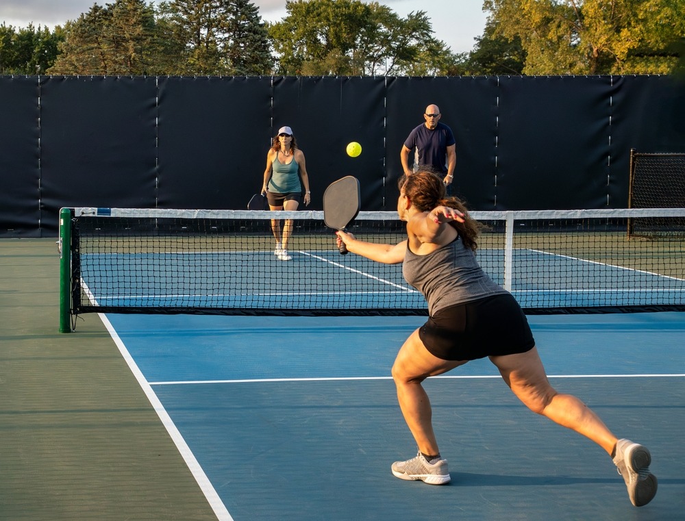 A woman lunges forward to hit a pickleball with a paddle on an outdoor court, while two other players stand ready on the opposite side of the net on a sunny day.