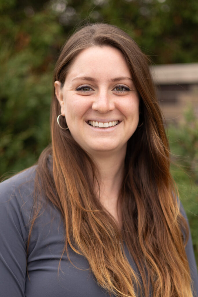 A woman with long brown hair and hoop earrings smiles outdoors, wearing a gray top, with greenery and a wooden structure blurred in the background.