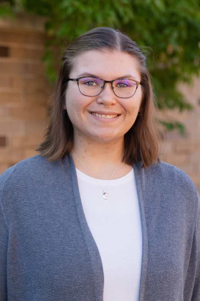 A young woman with shoulder-length brown hair and glasses smiles at the camera. She is wearing a white top, a gray cardigan, and a silver necklace. The background includes a brick wall and green foliage.
