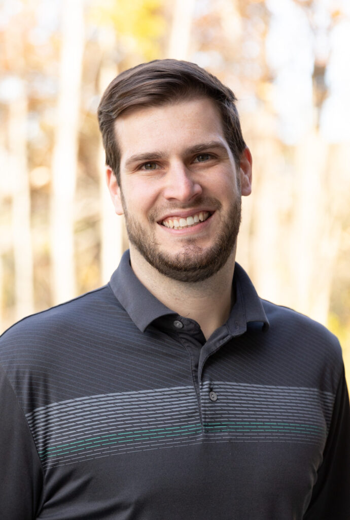 A man with short brown hair and a beard smiles outdoors. He is wearing a dark gray collared shirt with horizontal stripes. The background is softly blurred with trees and sunlight.