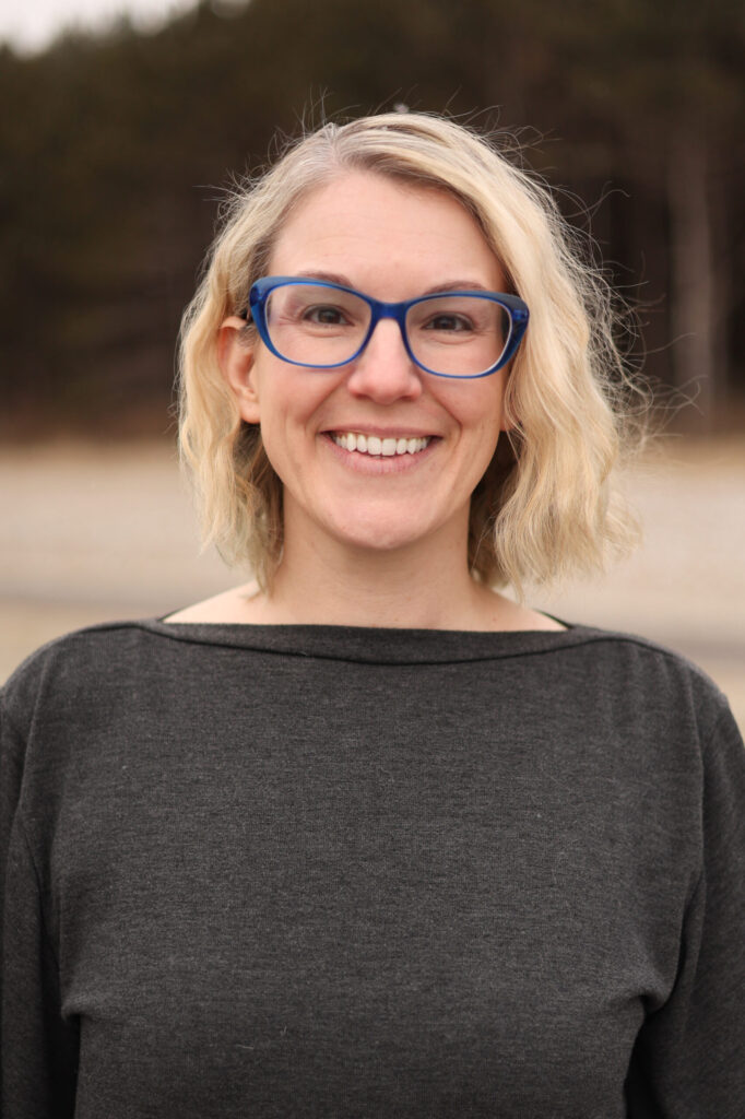 A woman with blonde wavy hair and blue glasses smiles at the camera. She is wearing a dark gray top and standing outdoors, with trees and a blurred background behind her.