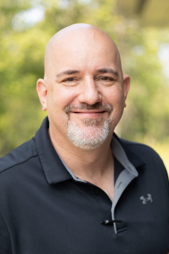 A bald man with a goatee smiles while wearing a black collared Under Armour shirt, standing outdoors with a blurred green background.