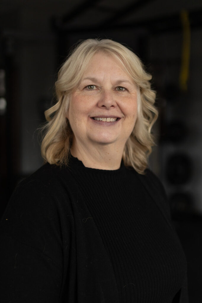 A smiling middle-aged woman with wavy blonde hair, wearing a black top and black cardigan, stands indoors with a dark, blurred background.