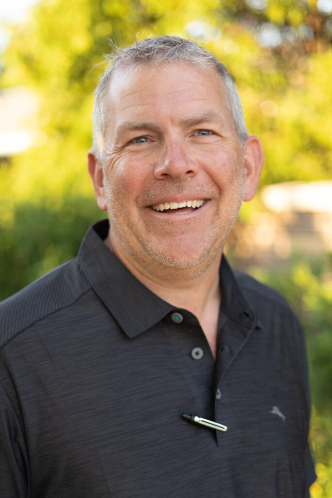 A middle-aged man with short gray hair and a stubbled beard smiles outdoors. He wears a dark collared shirt with a pen clipped to it, and the background is blurred greenery in sunlight.