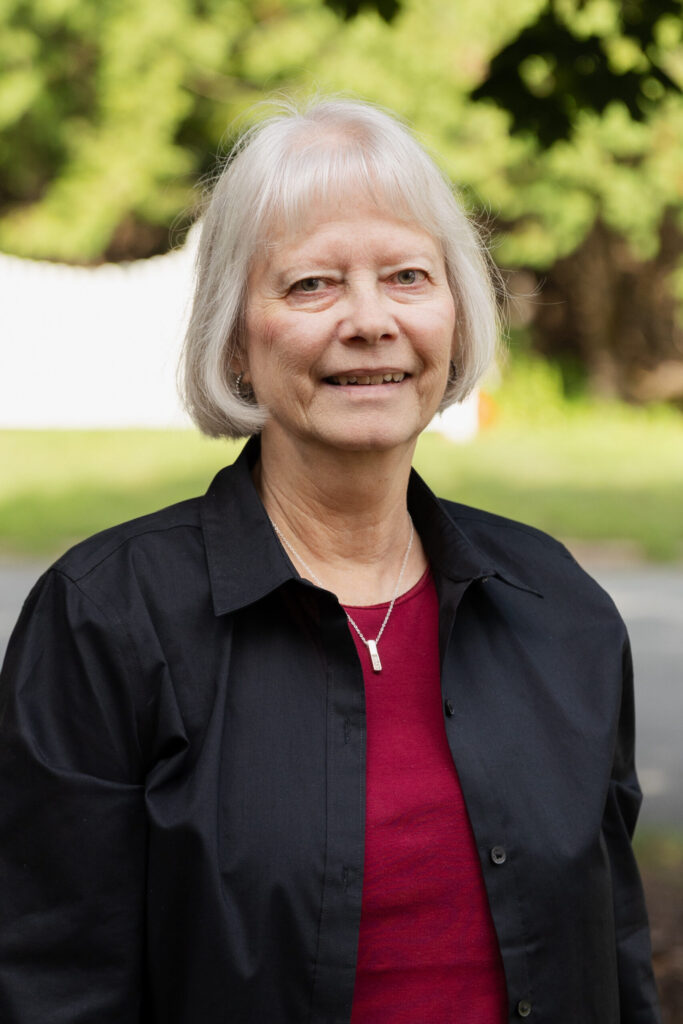 An older woman with short white hair, wearing a black shirt over a red top and a silver necklace, stands outdoors with greenery and a blurred background behind her.