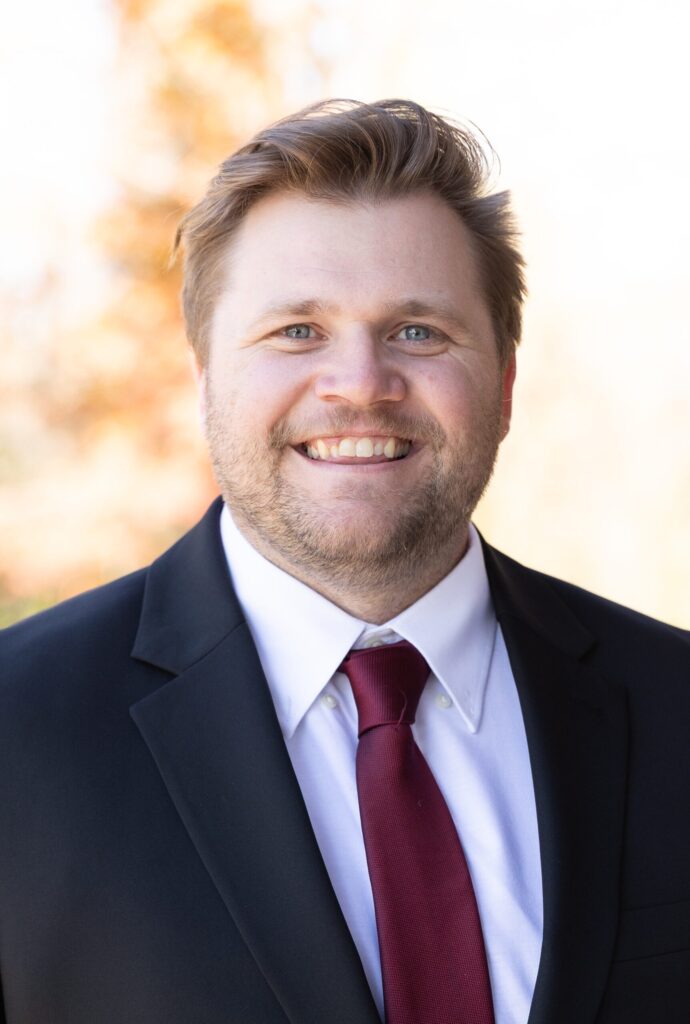 A smiling man with light hair and a beard wears a black suit, white shirt, and dark red tie. The background is softly blurred with warm, natural colors.
