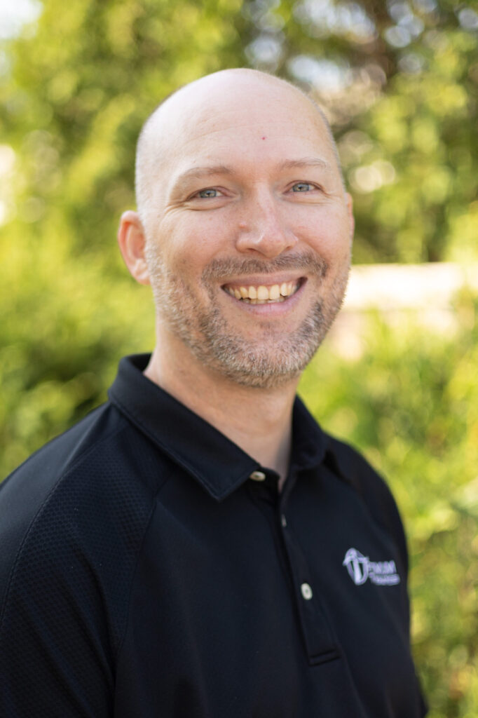A smiling, bald man with a short beard is wearing a black collared shirt and standing outdoors with green, blurry foliage in the background.