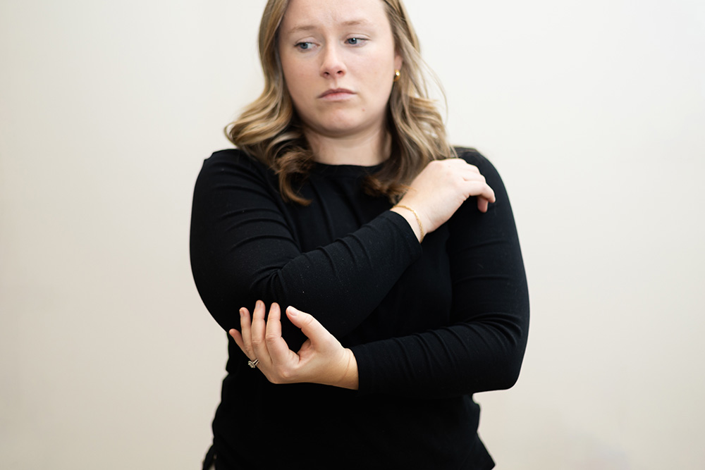 A woman with light brown hair, wearing a black long-sleeve shirt, looks concerned while holding and gently touching her left elbow with her right hand.