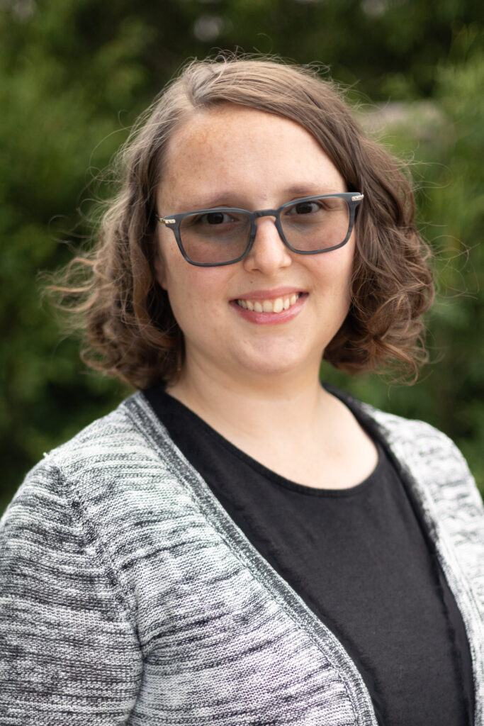 A woman with wavy brown hair and glasses smiles at the camera. She is wearing a black top with a gray patterned cardigan, and the background is blurred greenery.