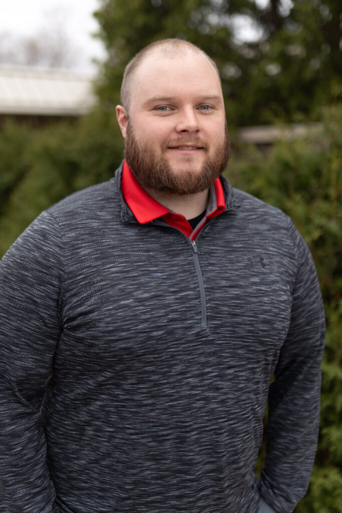 A bearded man with short hair is wearing a gray quarter-zip sweater over a red collared shirt, standing outdoors in front of green bushes and trees, smiling slightly at the camera.