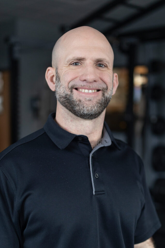 A smiling bald man with a trimmed beard wearing a black collared shirt stands indoors, with gym equipment blurred in the background.