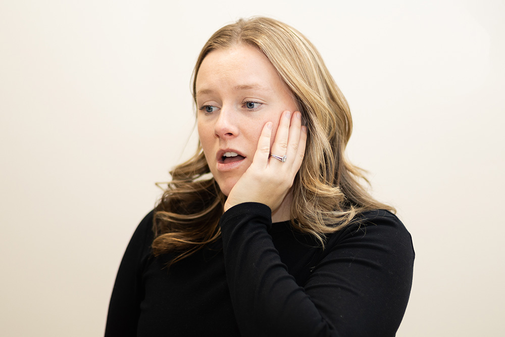 A woman with light brown hair wearing a black top holds her hand to her cheek, appearing to be in pain or discomfort, with her mouth slightly open and a concerned expression on her face.