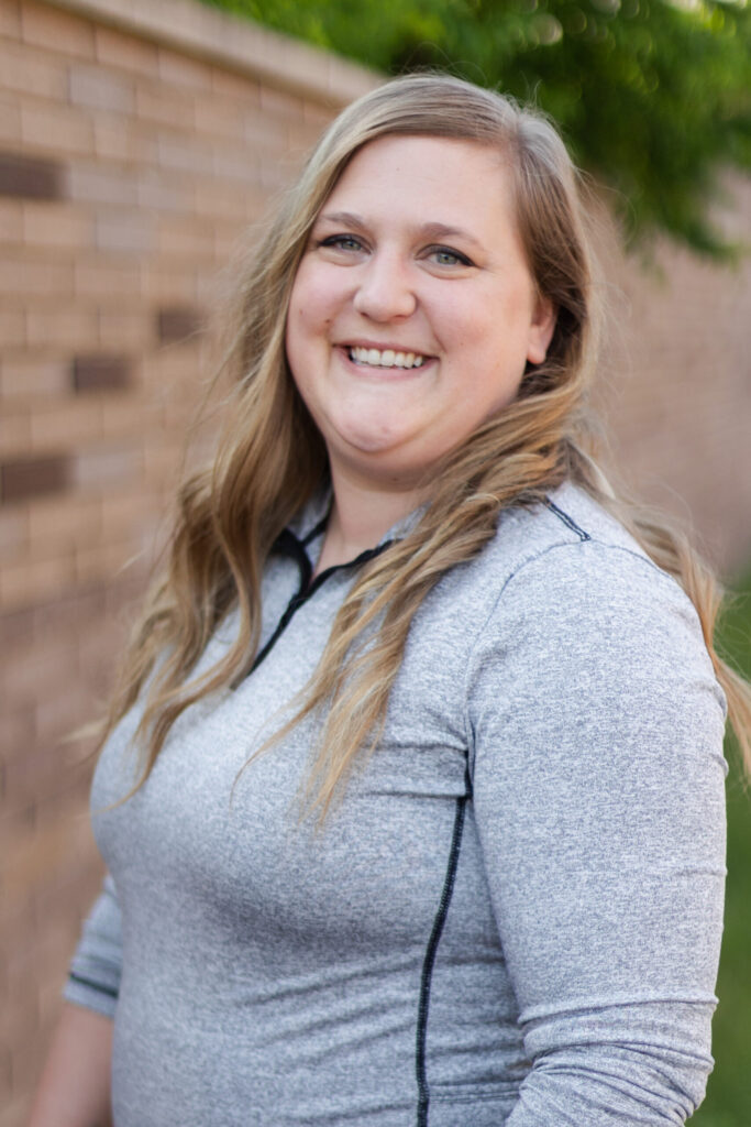 A woman with long blonde hair wearing a light gray zip-up shirt smiles while standing outdoors in front of a brick wall and greenery.