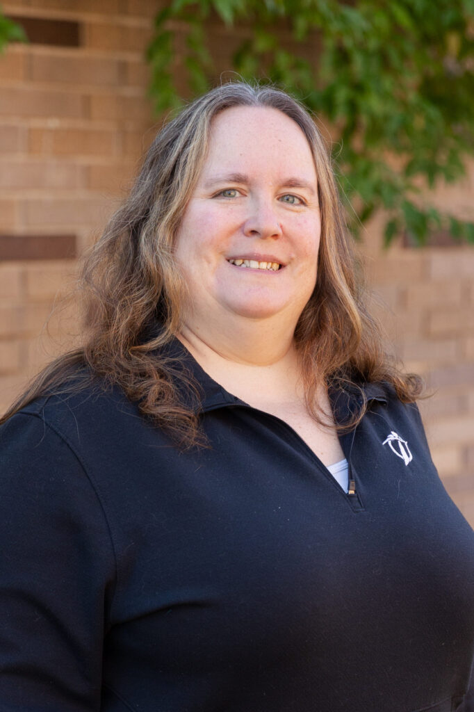 A woman with long, wavy brown hair and wearing a black zip-up top stands in front of a brick wall and green leafy branches, smiling at the camera.