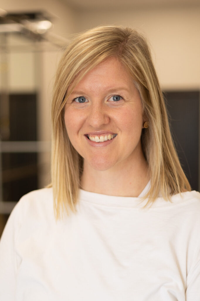 A woman with straight blonde hair and blue eyes smiles at the camera. She is wearing a white top and is standing indoors with a blurred background.