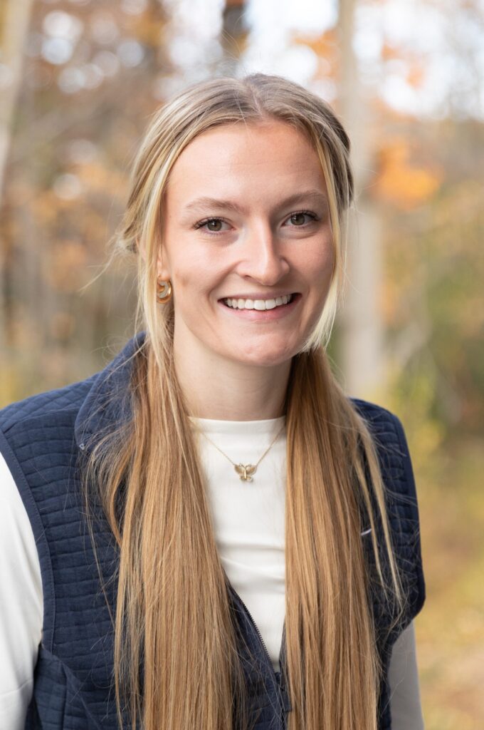 A young woman with long blonde hair, wearing a navy vest over a white shirt, stands outdoors smiling. Autumn trees with yellow and brown leaves provide a soft, blurred background.