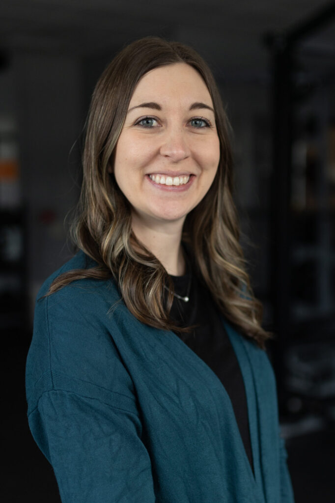 A woman with long brown hair, wearing a teal cardigan over a black top, smiles at the camera. The background is softly blurred, suggesting an indoor setting.