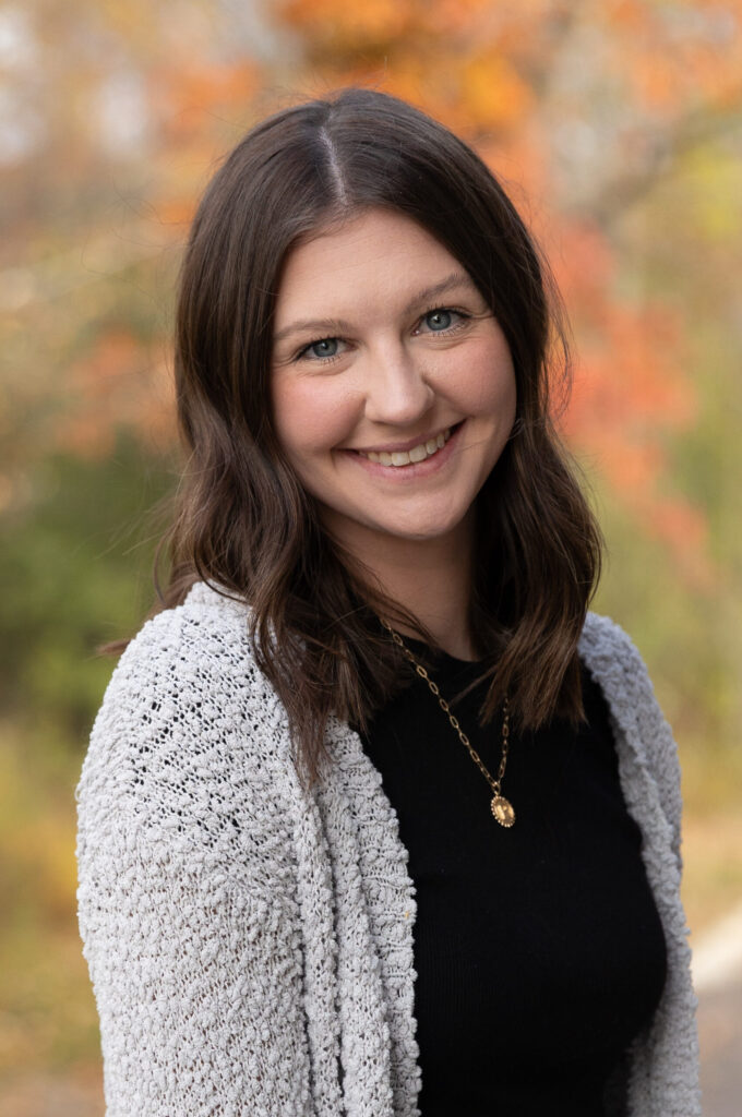 A woman with wavy brown hair, wearing a gray cardigan, black top, and gold necklace, smiles at the camera. The background is outdoors with blurred fall foliage in shades of orange and green.