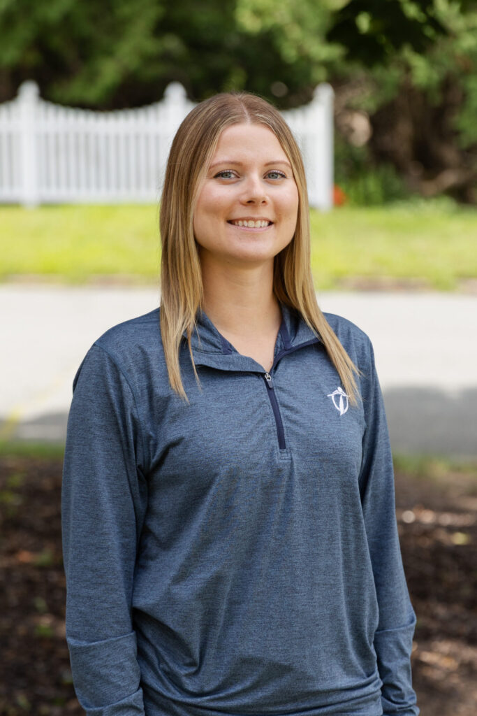 A young woman with straight blonde hair and a blue zip-up pullover stands outdoors, smiling. There is greenery and a white fence blurred in the background.
