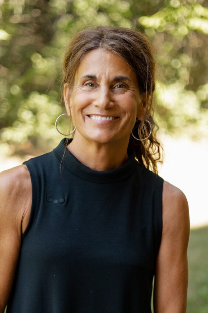 A woman with wavy brown hair tied back, wearing large hoop earrings and a sleeveless black top, smiles while standing outdoors with greenery in the blurred background.