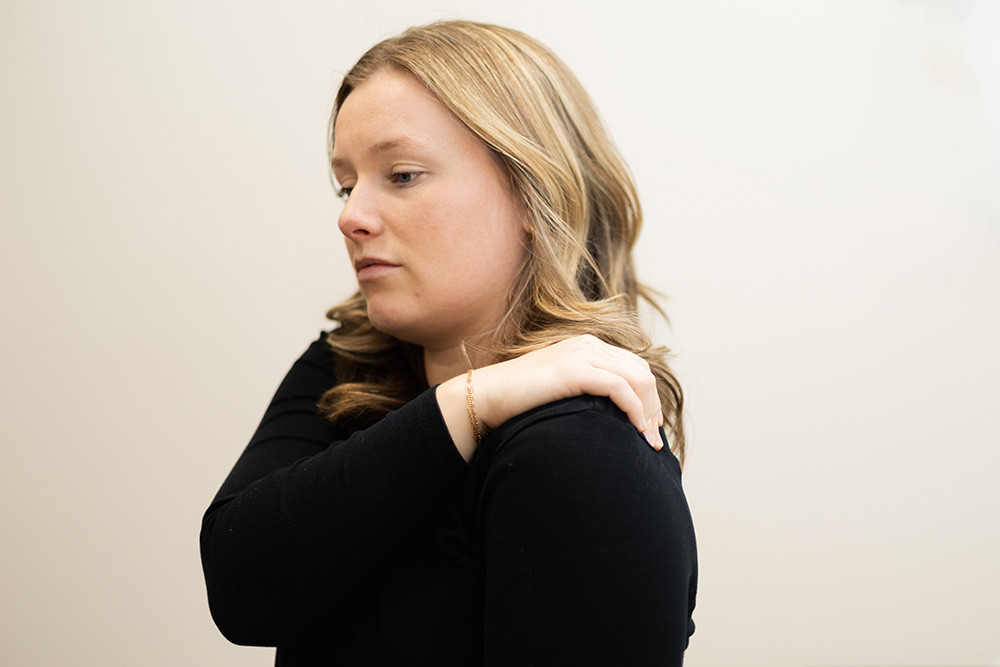 A woman with light brown hair wearing a black top holds her left hand on her right shoulder, appearing to feel discomfort or pain. The background is plain and light-colored.