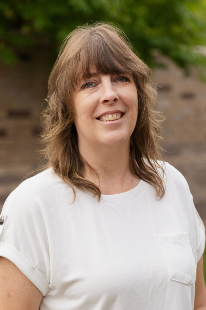 A woman with wavy light brown hair and bangs, wearing a white short-sleeved top, smiles while standing outdoors in front of a blurred brick wall and green foliage.