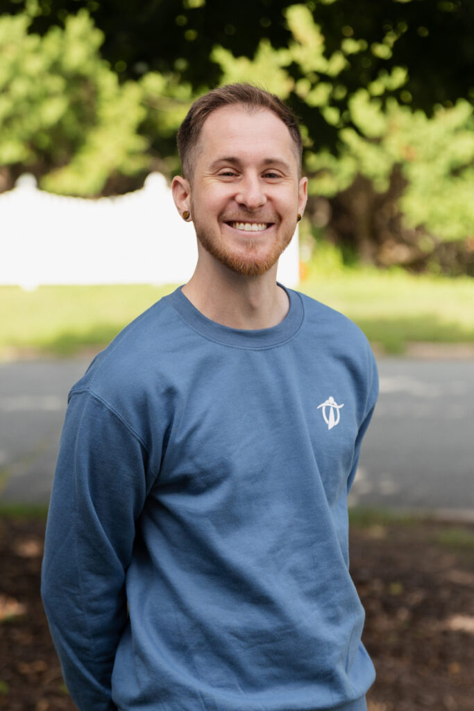 A man with short light brown hair and a beard is smiling outdoors, wearing a blue sweatshirt. Green trees and a blurred street are visible in the background.
