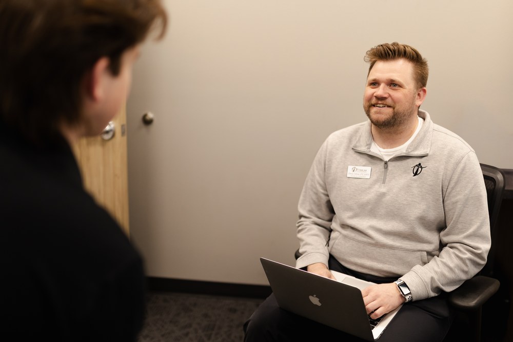 A man with a laptop sits and smiles while talking to another person in an office setting. He wears a light gray sweater, name tag, and a smartwatch. The other person is mostly out of focus in the foreground.