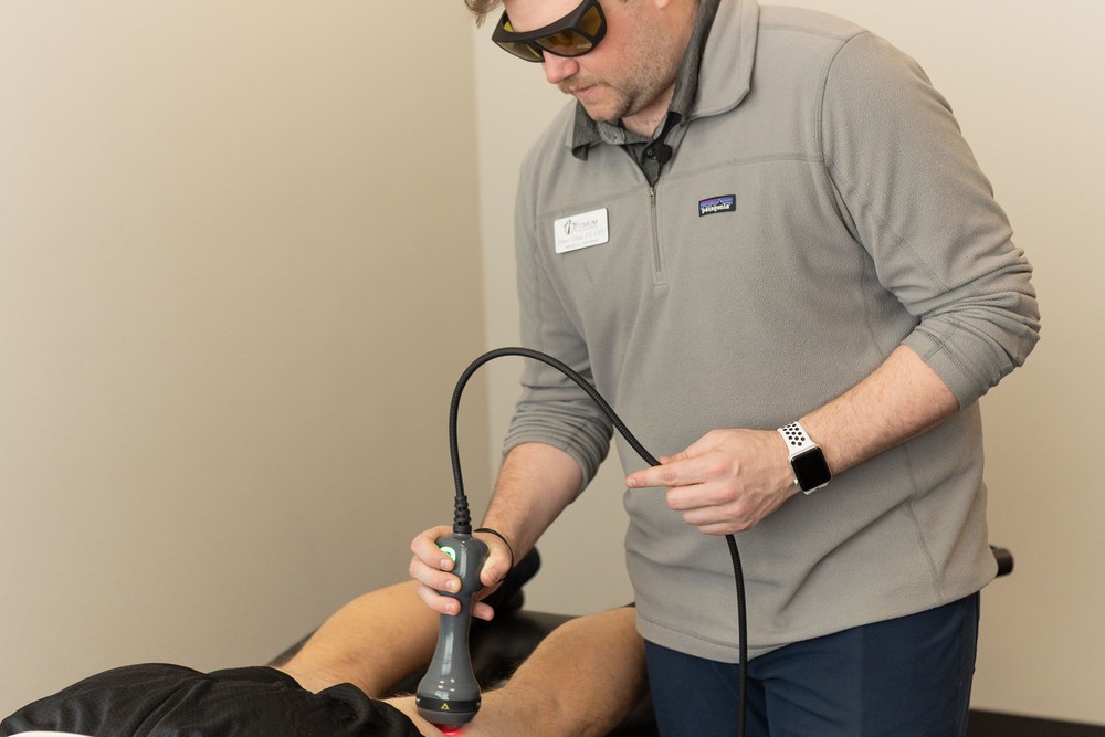 A person wearing glasses uses a handheld device to perform a treatment on another persons leg, who is lying down and covered with a black cloth. The setting appears to be a clinical or therapy room.