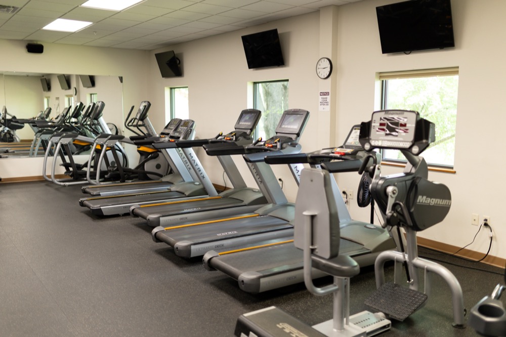 An empty gym features a row of gray Matrix treadmills, a Magnum exercise machine in the foreground, windows, wall-mounted TVs, and a large mirror reflecting the equipment.