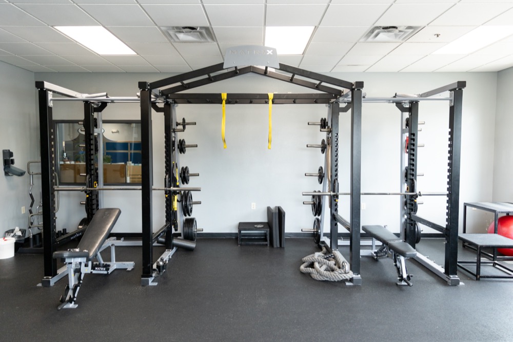 A gym room with weight racks, benches, yellow suspension straps, medicine balls, battle ropes, and mirrors, all set on a black rubber floor under fluorescent lights.