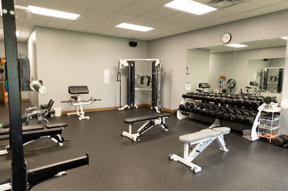 A gym room with benches, a cable machine, dumbbells on a rack, a large mirror, and a wall clock above. The floor is rubberized and the walls are light gray.