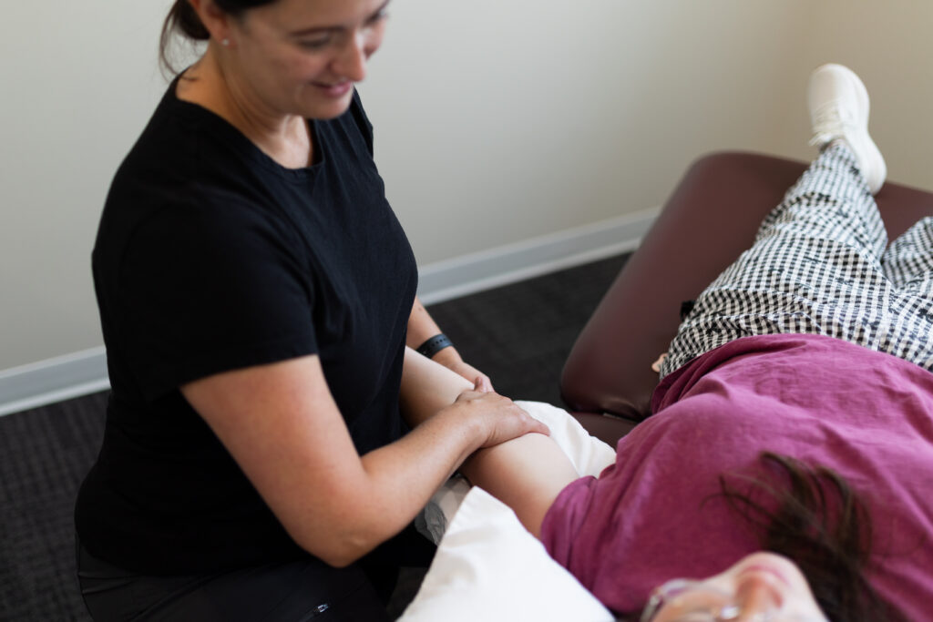 A woman in black holds and supports the arm of a person lying on a table, who is wearing a pink shirt and patterned pants, during a physical therapy session.