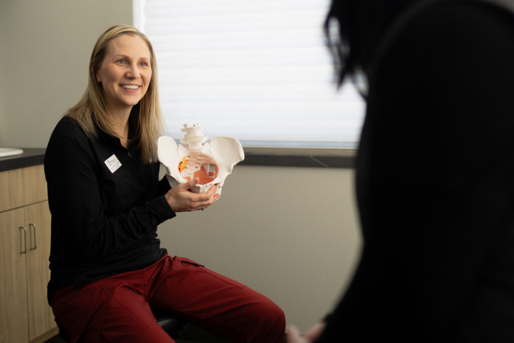 A woman in red pants smiles while holding a pelvic bone model, speaking to another person in a medical office.