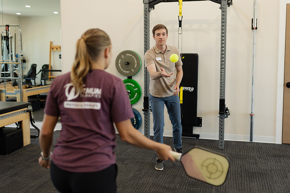 A man tosses a yellow ball toward a woman holding a pickleball paddle in a gym or physical therapy room with exercise equipment visible in the background.